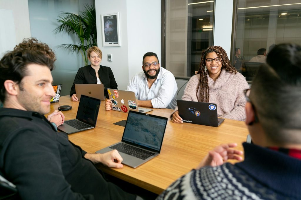 About Us 1 four people all on laptops, two men and two women, listen to person talking in a board meeting